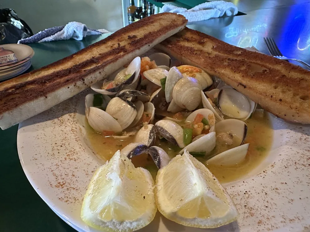 A plate of steamed clams in a broth with herbs and vegetables, served with two long slices of toasted bread and lemon wedges on a white plate.