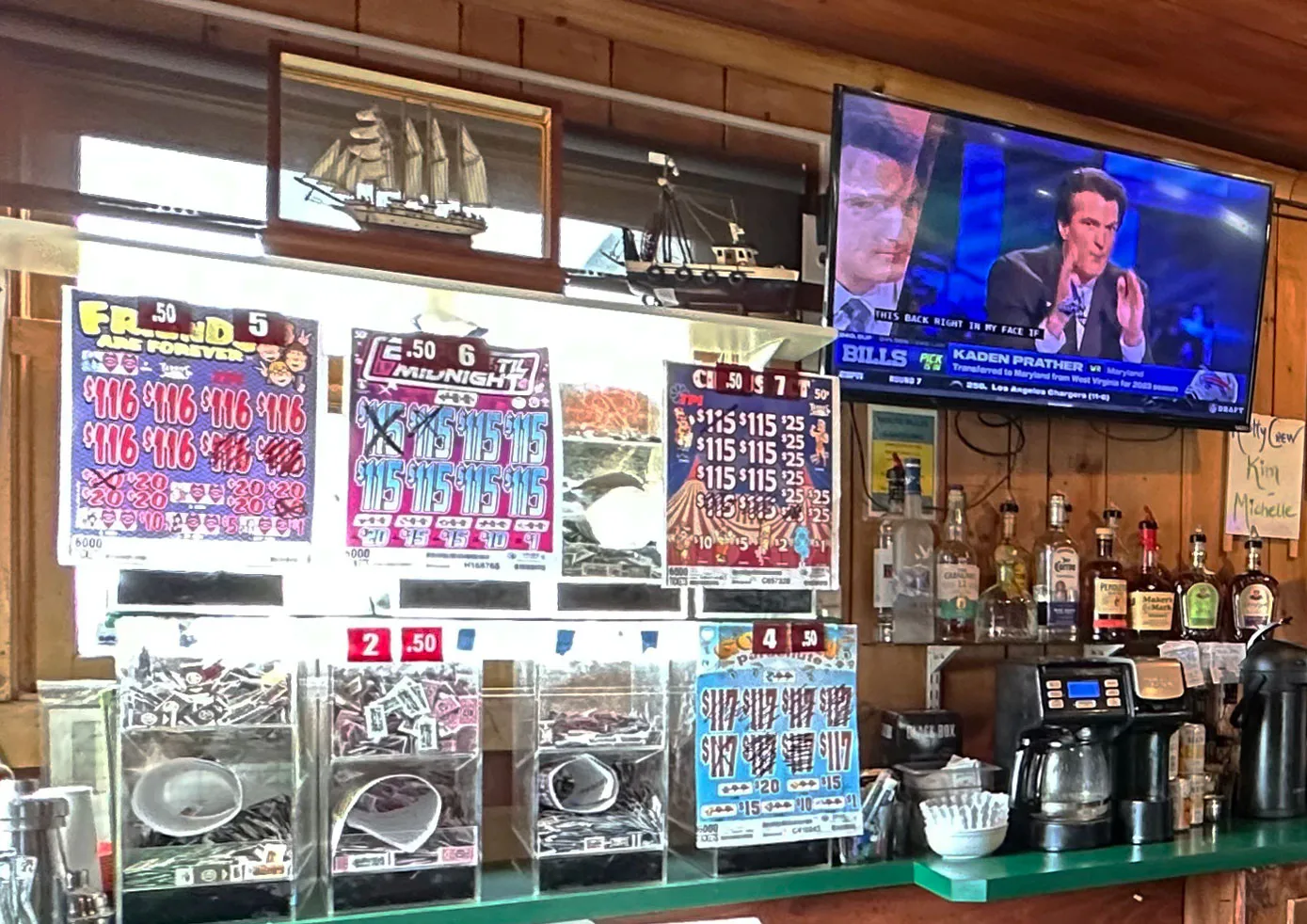 Bar with pull-tab lottery tickets displayed below a TV showing a sports commentator. The counter has liquor bottles and a coffee maker, with nautical decor above.