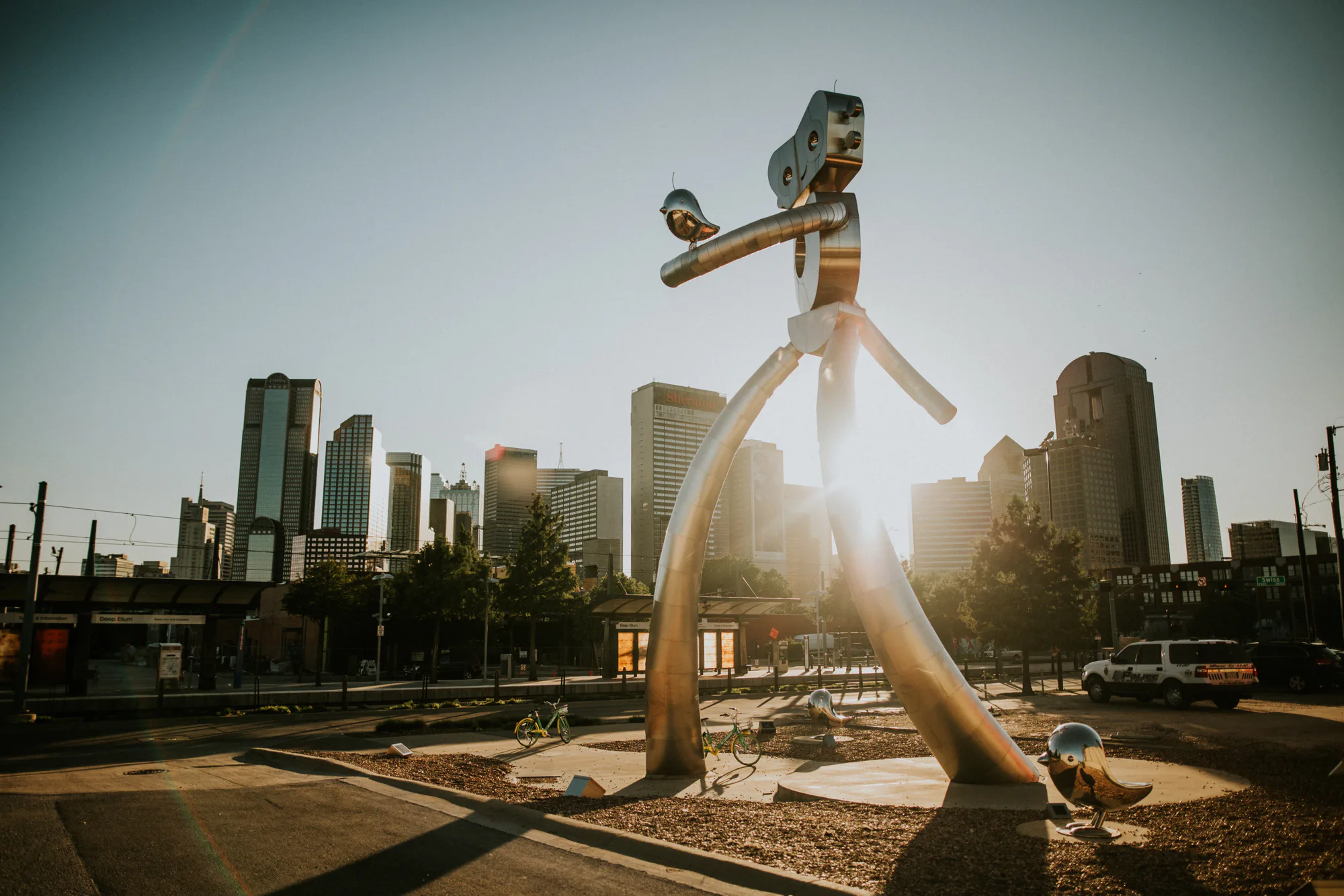 Large shiny metal stick-figure sculpture with a small bird perched on its arm, city skyline at sunrise