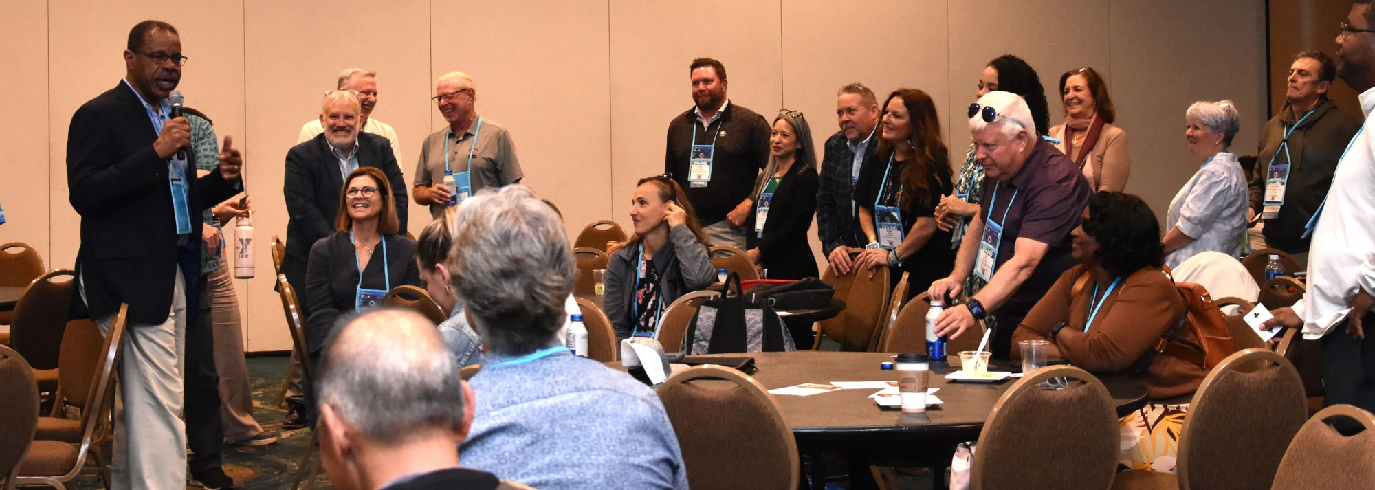 Group of conference attendees in a meeting room listening as a man speaks into a microphone
