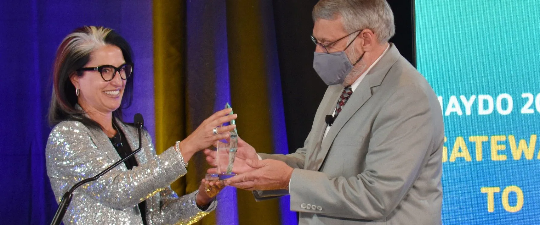 A woman in a glittery jacket and glasses smiles while accepting an award from a man in a light gray suit and mask. Background screen displays NAYDO 2021 GATEWAY TO GIVING.