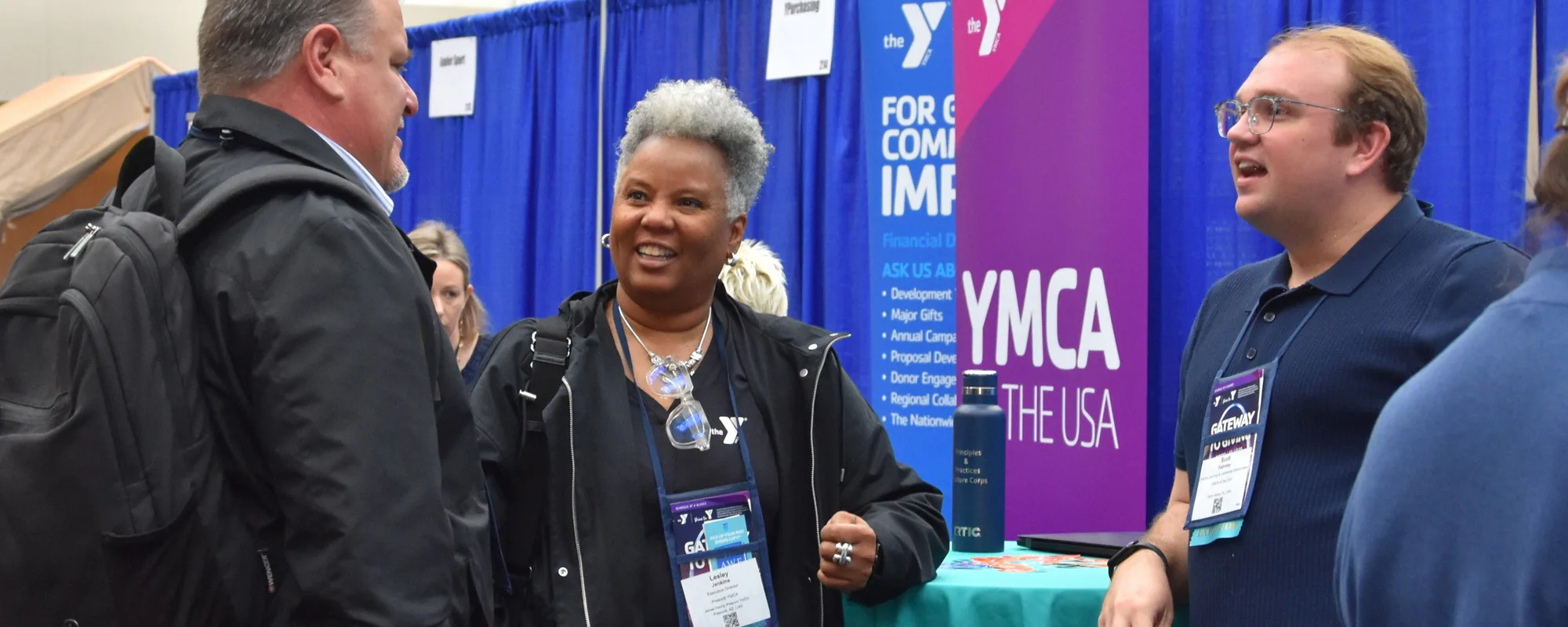 Three people engage in conversation at a YMCA event booth, with informational banners in the background. One person wears a backpack and another holds a water bottle.
