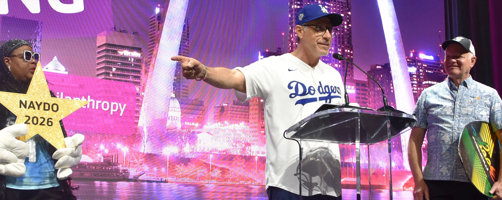 A person wearing a Dodgers jersey speaks at a podium. Another holds a yellow star with NAYDO 2026. A third person stands beside them. A city skyline is in the background.