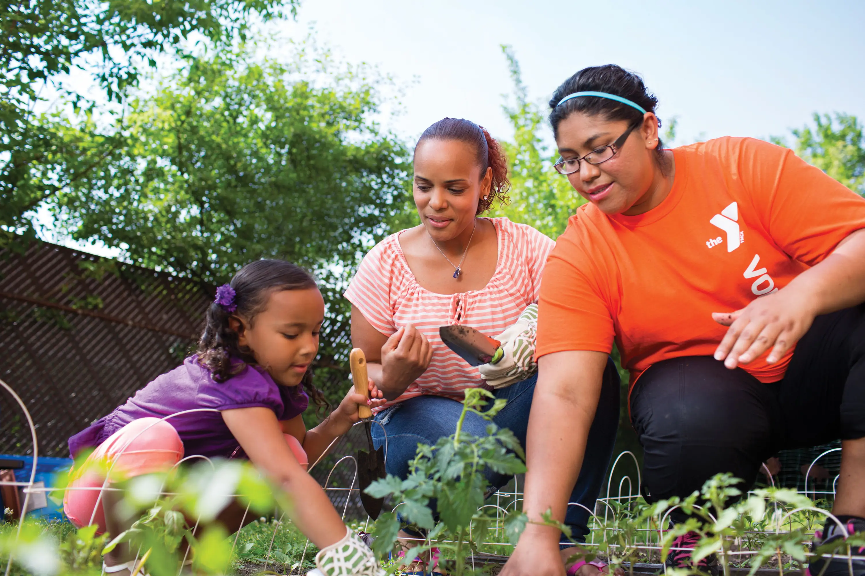 A young girl, a woman, and a volunteer in an orange shirt are gardening together, tending to plants in a garden. They are surrounded by greenery and bright sunlight.