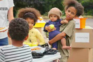 Children gather near a table with food and donation boxes, one holding a toddler with an apple. A volunteer shirt is partially visible on an adult nearby.