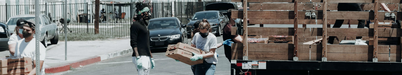 People wearing masks and gloves are unloading food boxes from a truck on a city street, engaged in a community distribution effort. Cars are parked in the background.