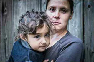 A woman holding a young child close to her chest. The child has curly hair and wears a dark shirt, while the woman looks directly at the camera. A wooden fence is in the background.