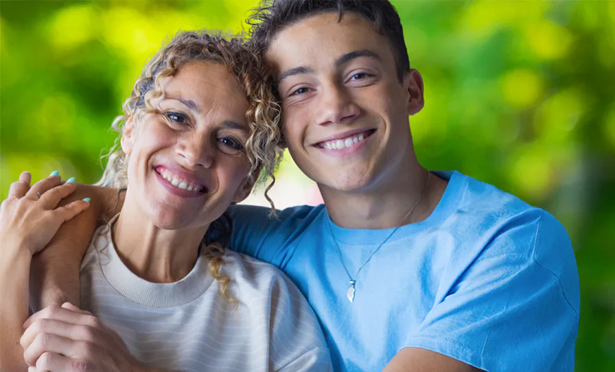 A woman and a young man stand closely, smiling warmly against a blurred green background. The woman has curly hair and wears a light sweater, while the man wears a blue shirt.