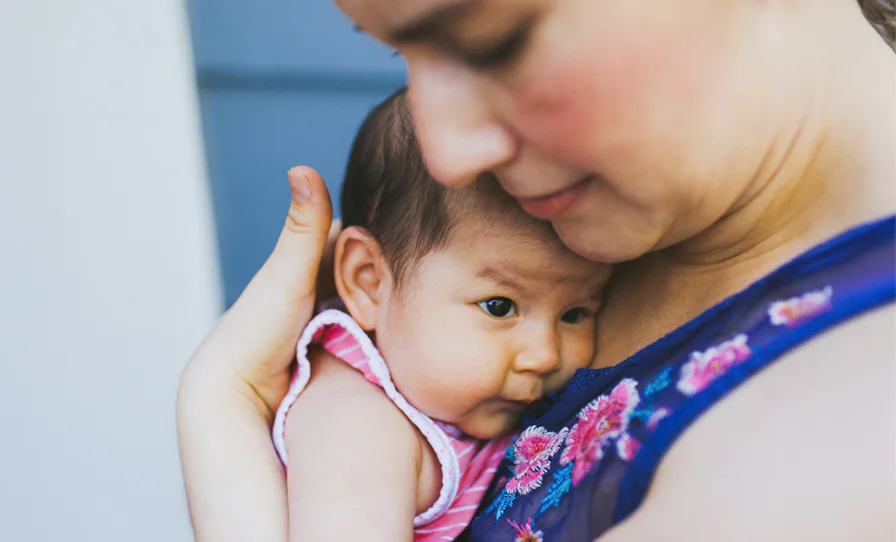 A woman gently holds a baby close to her chest, both wearing colorful clothing. The baby looks content, nestled against the woman's shoulder in a tender moment.