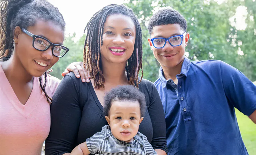 A smiling woman stands outdoors with three children, holding an infant in her arms. Two older kids, wearing glasses, stand close on either side. Trees are in the background.