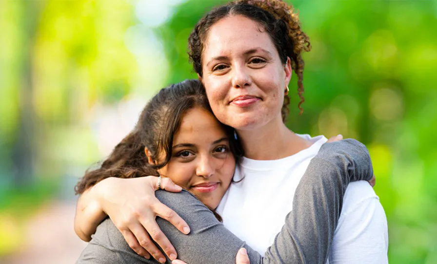 A woman and a girl share a warm embrace outdoors, both smiling gently. The background is a soft-focus green, suggesting a natural setting like a park or garden.