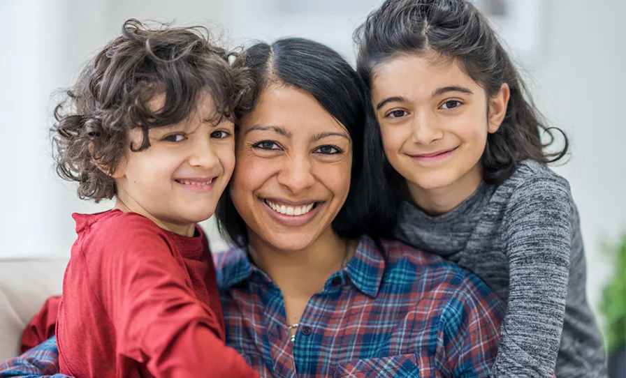 A woman smiles happily with two young children, one wearing a red shirt and the other in a gray top. They are close together in a bright, cozy setting.