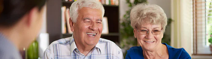 Elderly couple sitting together at a table, smiling and engaged in conversation with an unseen person across from them. A laptop is open on the table in a cozy, well-lit room.