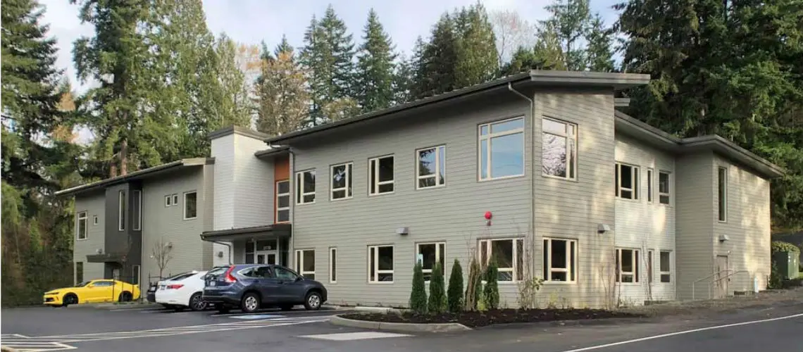 Modern two-story building with beige siding nestled among tall trees. Several cars parked in front including a yellow car. Large windows and a sloped roof are visible.