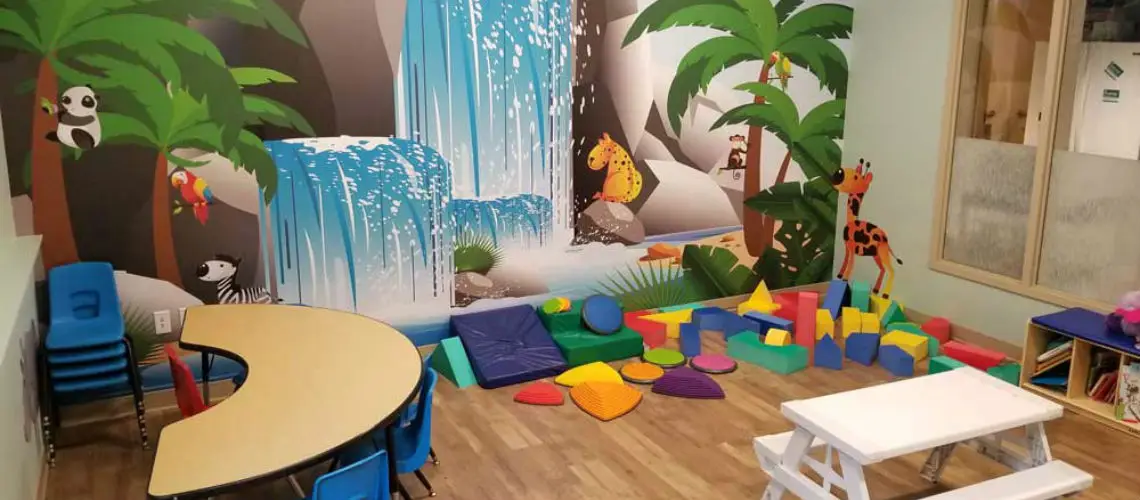 Children's playroom with mural of jungle animals and waterfall. Room has tables, chairs, and soft play blocks in various shapes and colors on a wood floor.