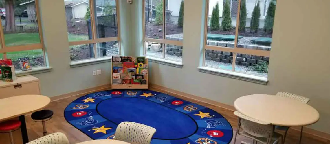 A bright classroom with large windows, featuring a colorful rug with stars, two round tables with chairs, and a book rack filled with various children’s books in the corner.