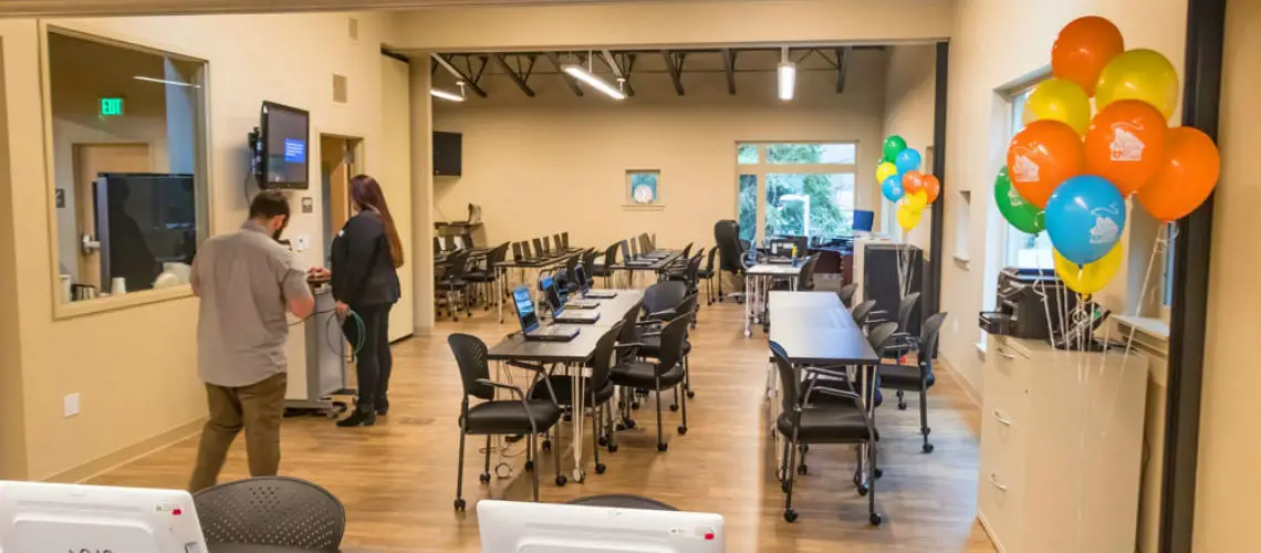 A large classroom with rows of tables and chairs, equipped with computers. Two people are setting up, and bright balloons are on the right. Natural light comes through the window.