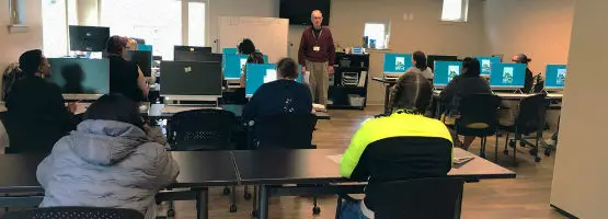 A classroom with students sitting at desks, each with a computer in front of them. A person stands at the front of the room, speaking to the class.