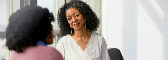 A woman with curly hair wearing a white top is sitting and attentively listening to another person in a pink top, in a brightly lit room with large windows.