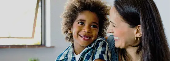 A child in a plaid shirt smiles joyfully at a woman beside them, who is smiling back warmly. They are indoors with a window in the background.