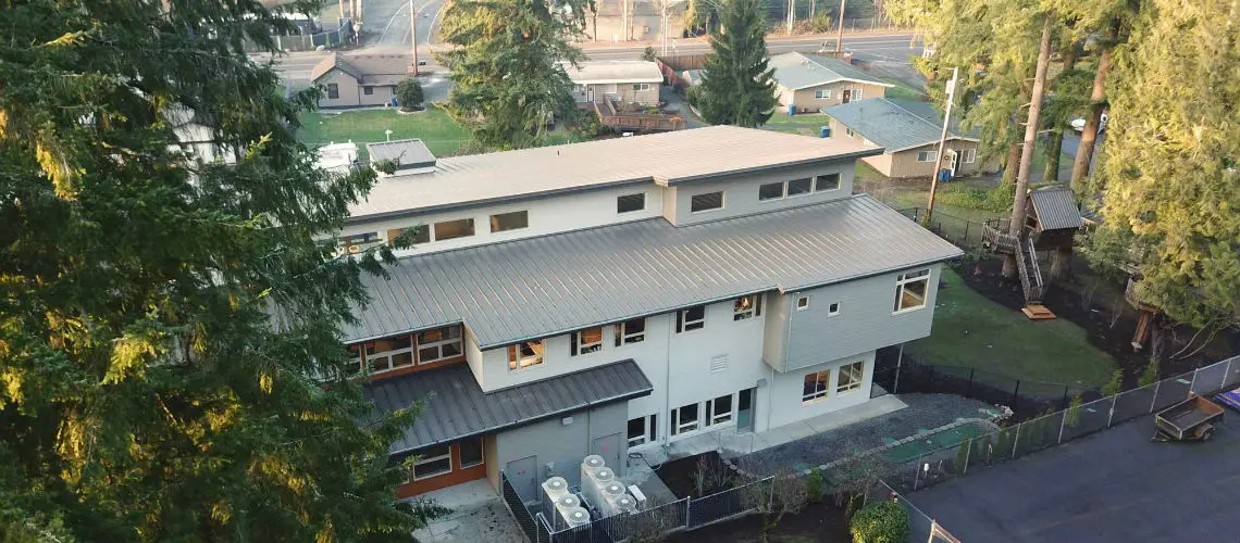 Aerial view of a modern two-story building with a metal roof, surrounded by trees. The building has multiple windows and is near a fenced yard with a path and greenery.