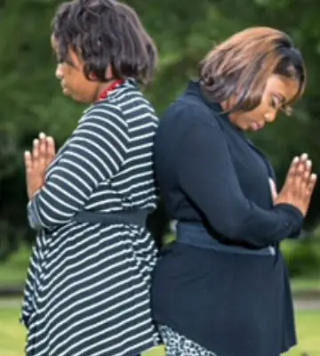 Two members standing back to back outdoors, eyes closed, hands together in prayer